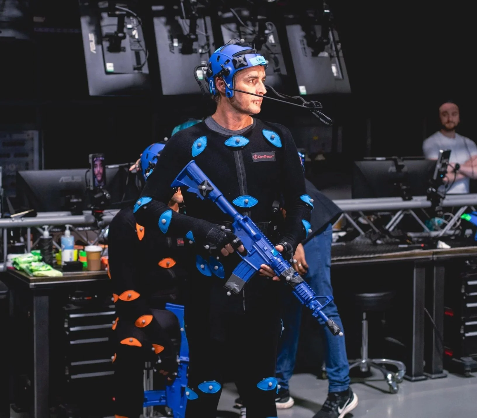 A man wearing motion capture gear stands in a motion capture studio with multiple screens and technicians in the background.