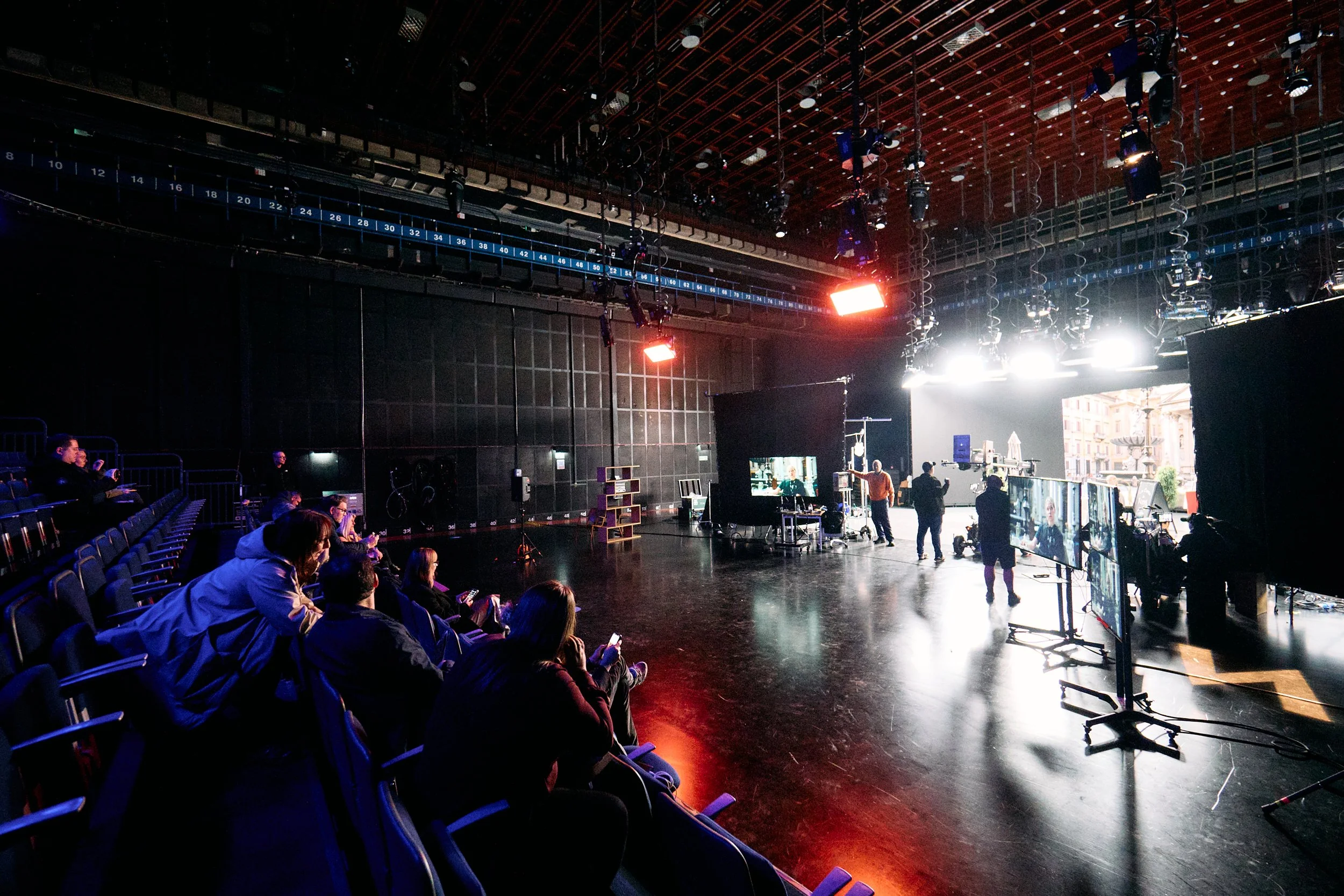 Interior view of a theater or studio with people seated and some standing, stage area with bright lights, various equipment, and large black curtains.