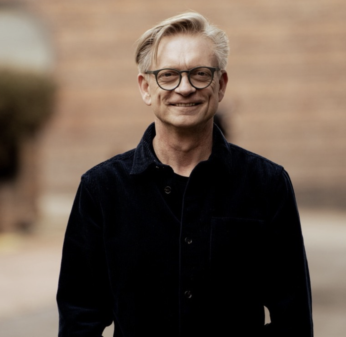 A middle-aged man with blonde hair, glasses, and a slight smile, wearing a black shirt, standing outdoors against a blurred brick wall background.