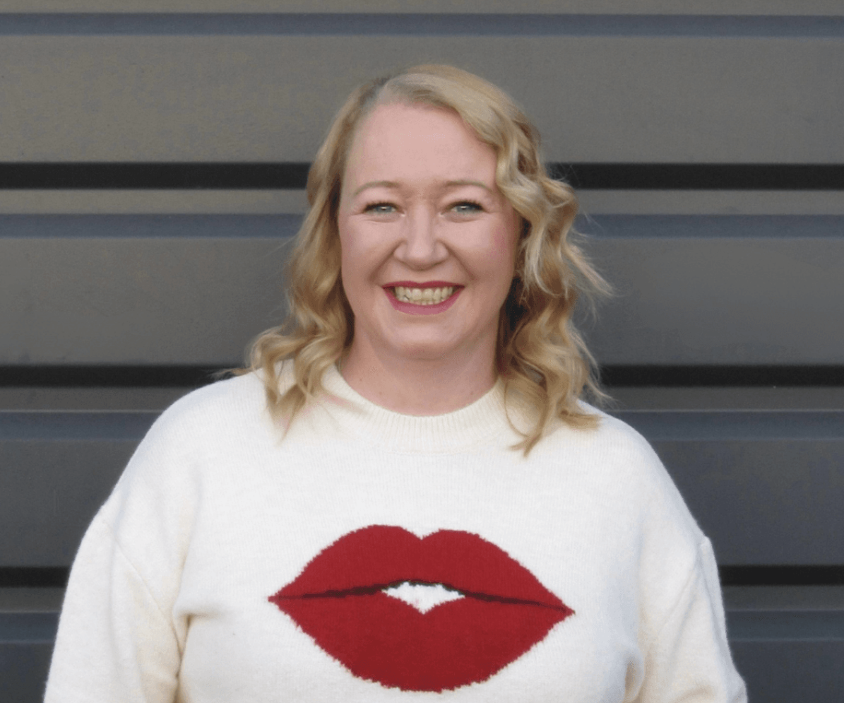 A smiling woman with wavy blonde hair wearing a white sweater with a red lip print on the front.