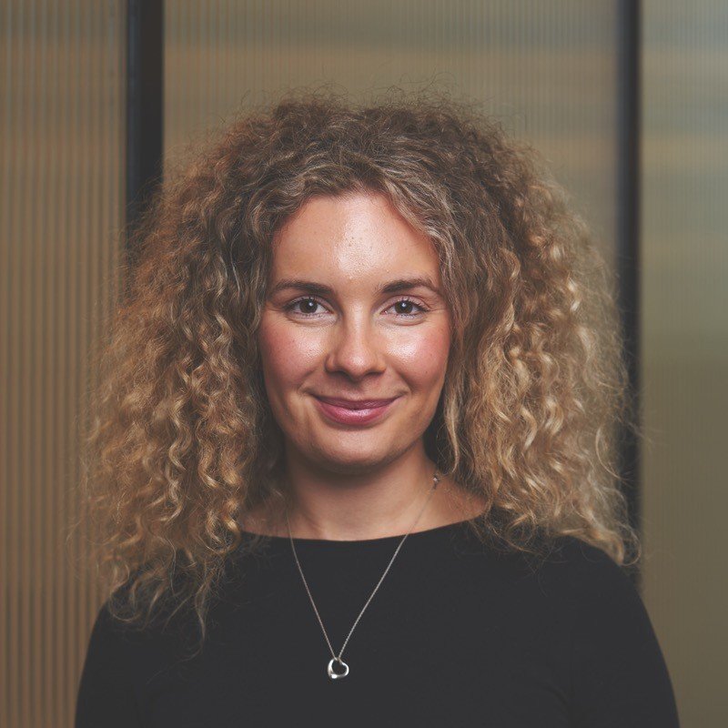 A woman with curly blonde hair smiling at the camera, wearing a black top and a silver heart-shaped necklace, standing indoors against a blurred background.