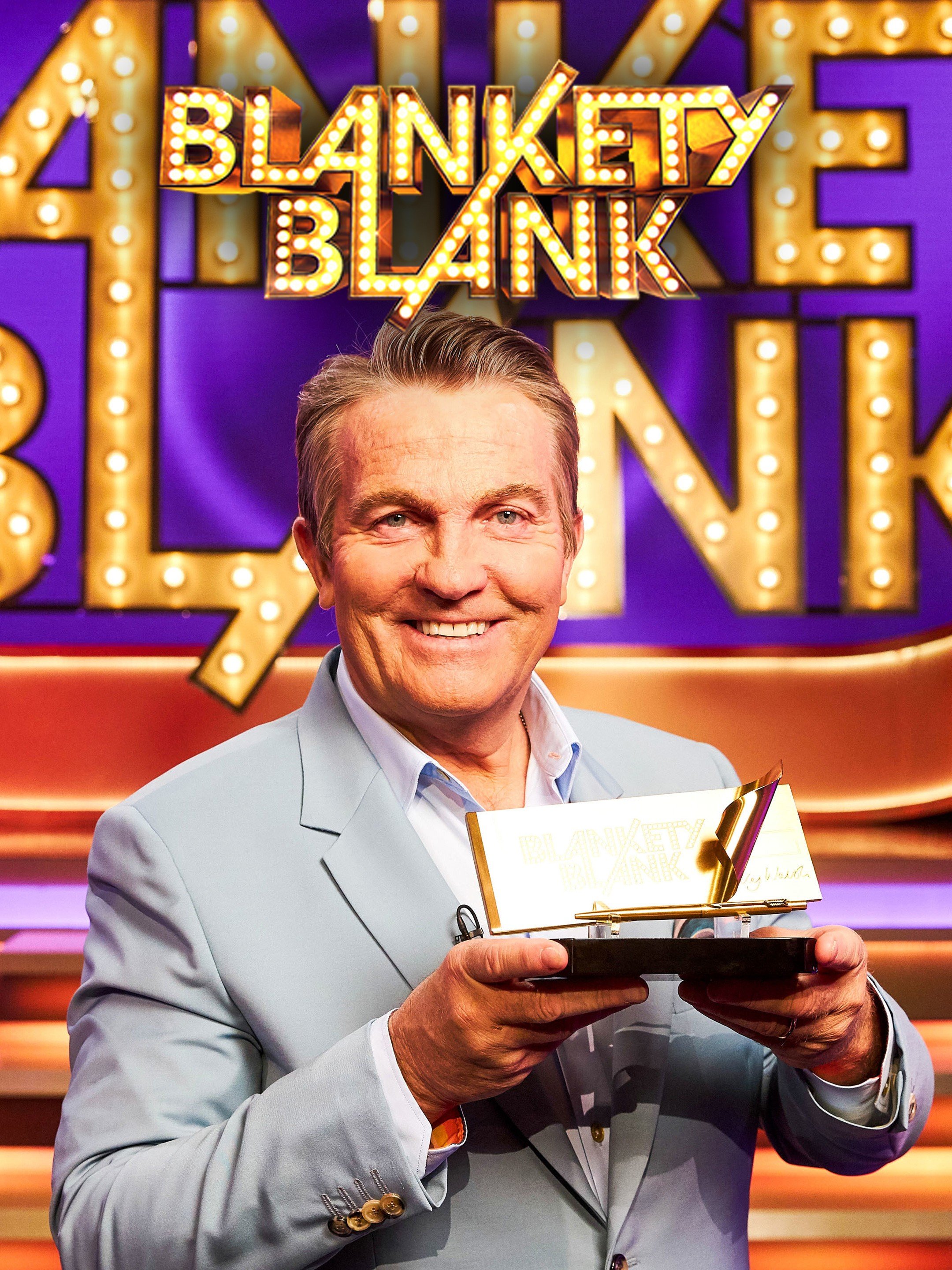 A man in a light gray suit holding a small trophy with the logo 'Blankety Blank' in bright lights, standing in front of a colorful, illuminated 'Blankety Blank' sign.