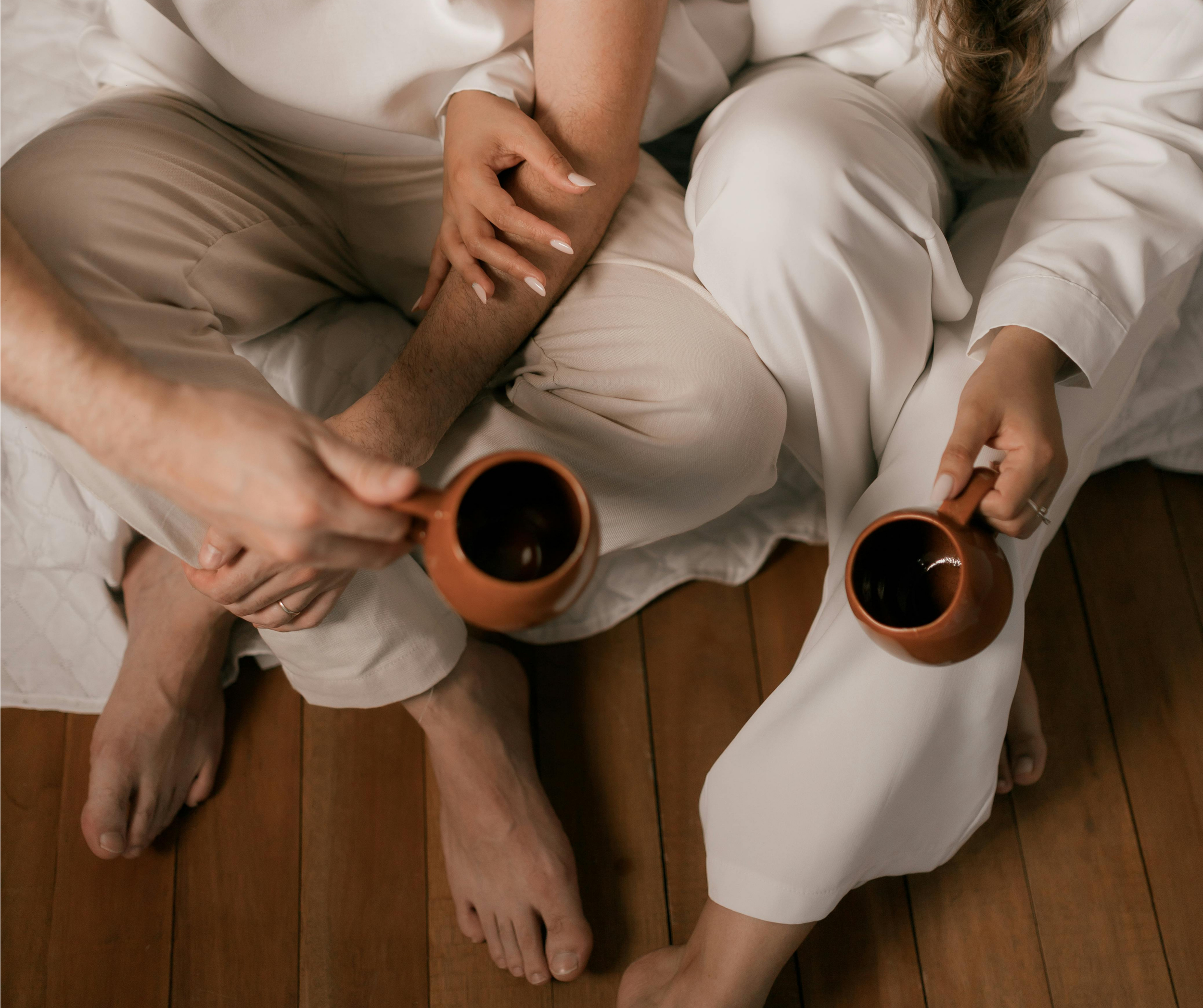 Twee mensen zitten op de grond en houden kopjes koffie vast. Ze dragen lichte, comfortabele kleding en hebben geen schoenen aan.