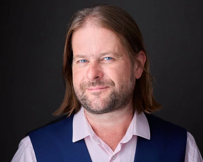 A middle-aged man with shoulder-length light brown hair and a beard, wearing a pink shirt and a dark vest, smiling slightly against a dark background.
