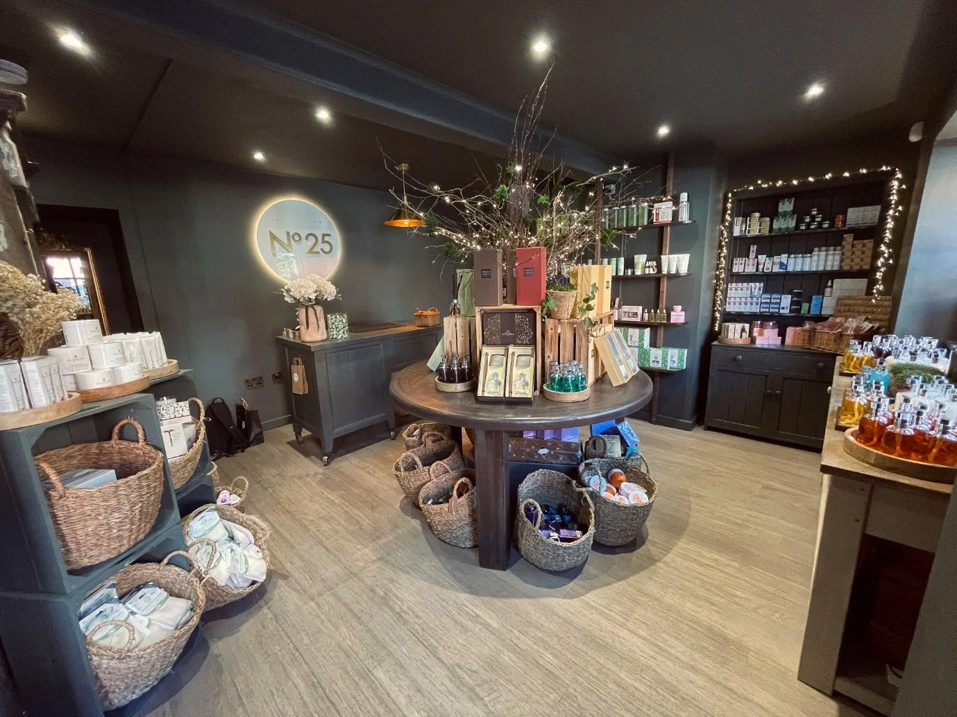 Interior of a retail store with shelves stocked with various bottles and packages, a central round table with baskets filled with products, and a wall with a lighted sign reading 'N° 25'.