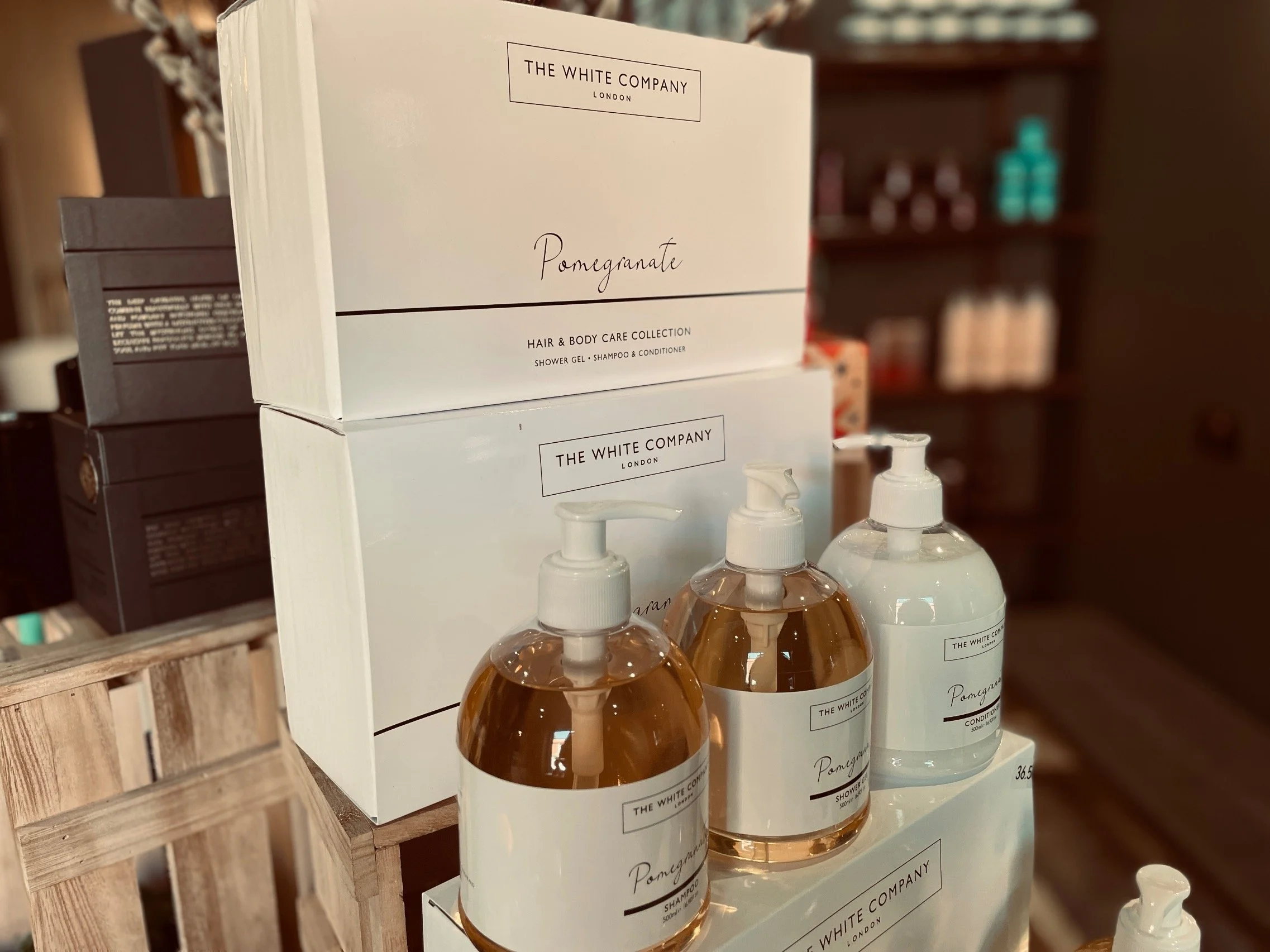White boxes and bottles of The White Company Pomegranate hair and body care collection, including shower gel, shampoo, and conditioner, on display on a wooden surface.