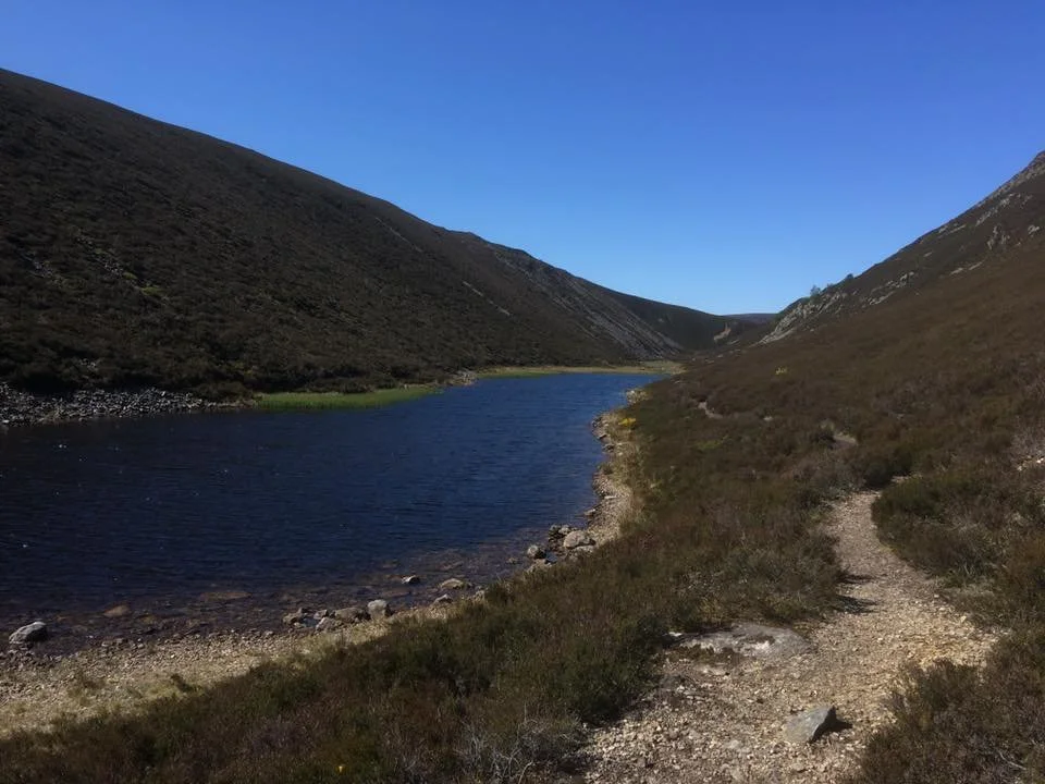 A mountain landscape with a narrow trail running alongside a deep blue lake, surrounded by brownish-green hills and a clear blue sky.