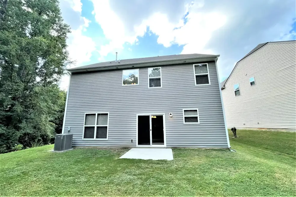 rear view of a two-story house with gray siding, a sliding glass door, and several windows, set on a grassy yard with trees and another house nearby.