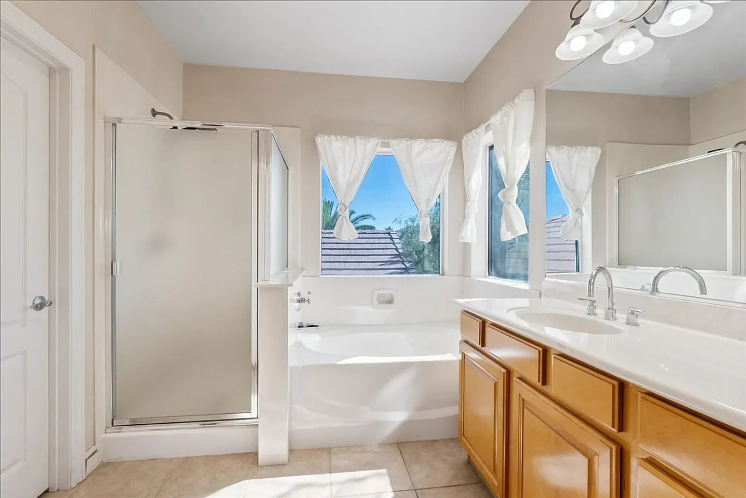 Bright bathroom with a window view of palm trees, featuring a bathtub, a separate shower stall, a large mirror, a wooden vanity, and white curtains.