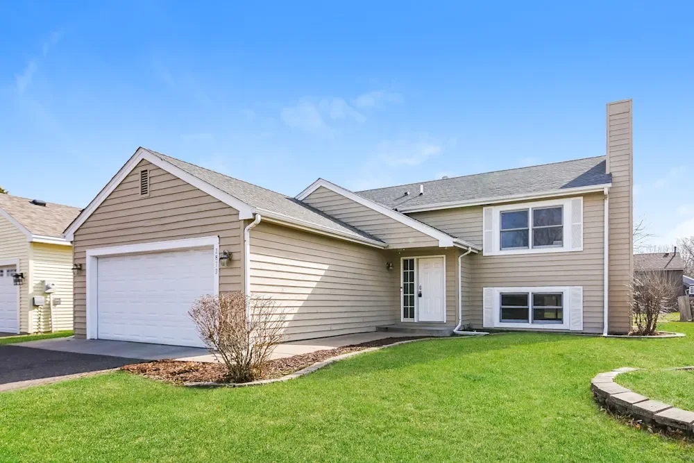 A beige two-story house with a white garage door and a well-maintained front lawn under a clear blue sky.