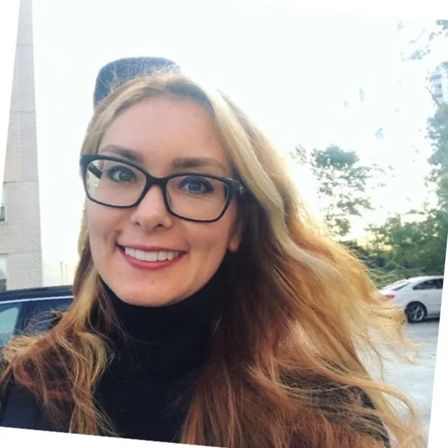 Smiling woman with glasses and long, wavy hair outdoors, with cars and trees in the background.