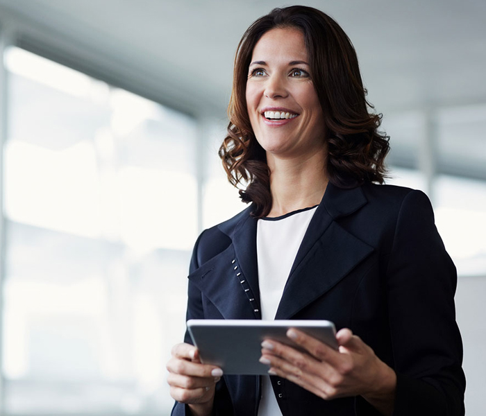 A smiling woman in business attire holding a tablet in an office setting with large windows.