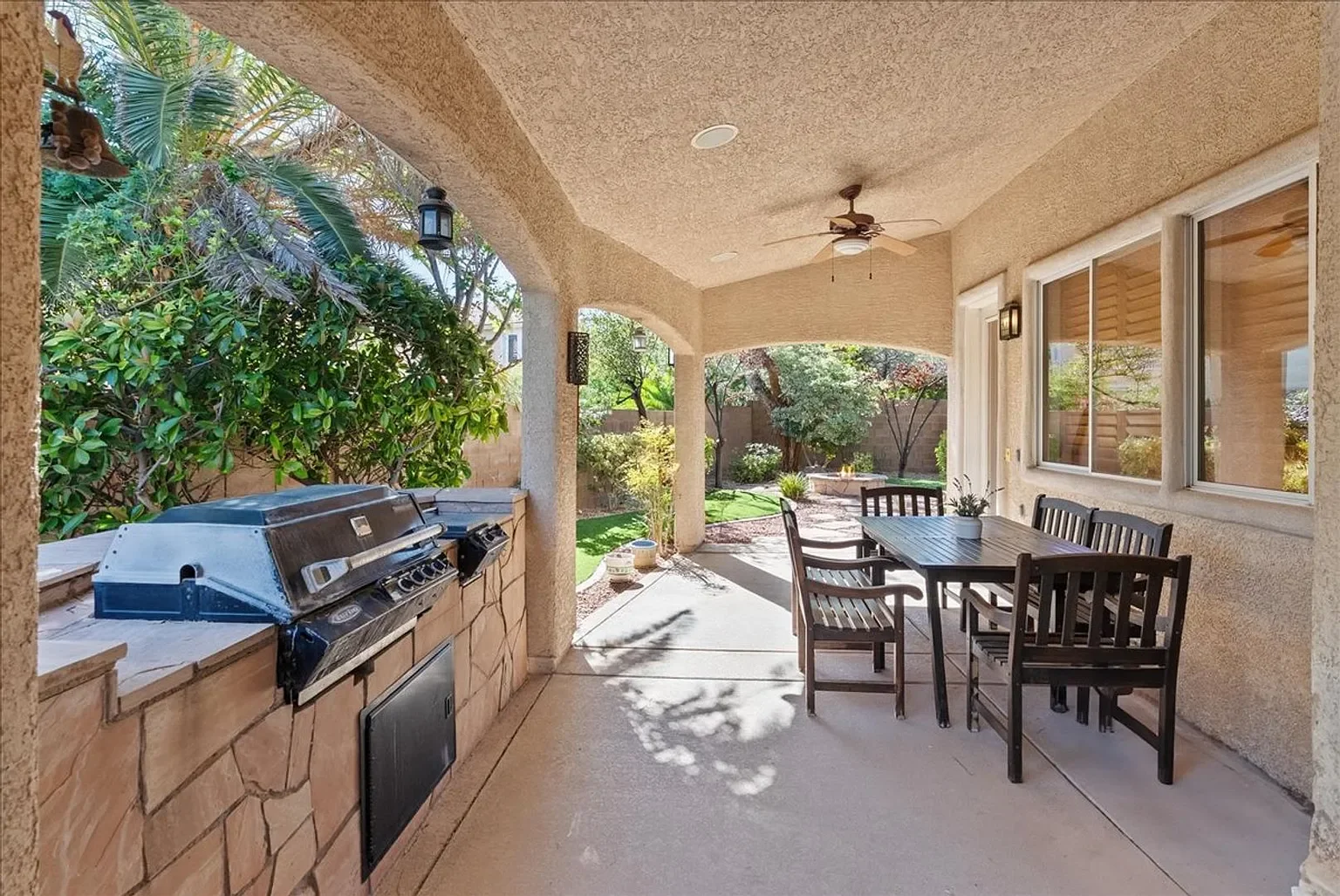 Covered backyard patio with outdoor grill, dining table with six chairs, ceiling fan, ceiling lights, and view of garden with trees and plants.
