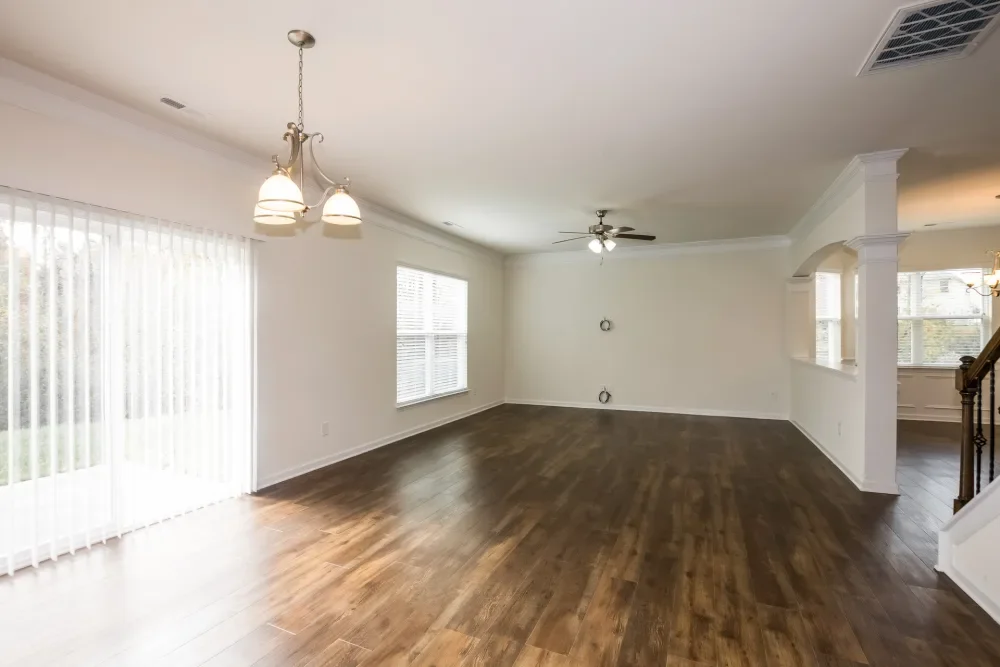 Empty living room with wooden floors, white walls, large windows with blinds, a ceiling fan, and a hanging light fixture.