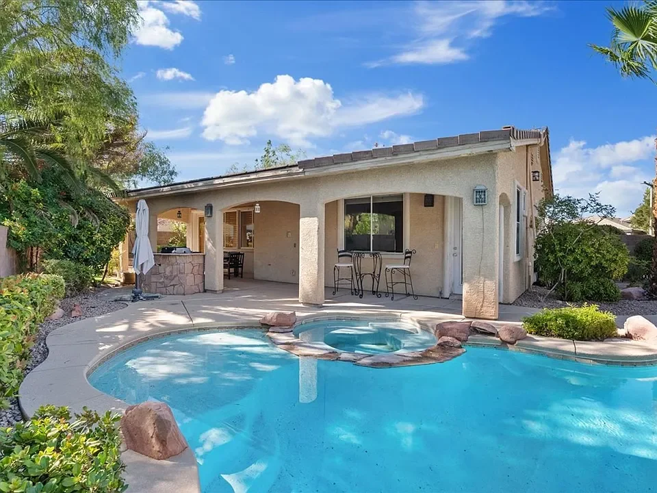 Backyard with swimming pool, hot tub, covered patio, outdoor seating, green bushes, trees, and a blue sky with clouds.