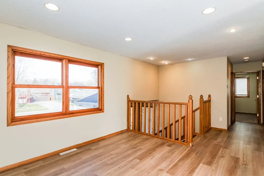 View of a bright, empty room with wooden floors, beige walls, a large window with wooden trim, and a wooden staircase with a railing leading down.