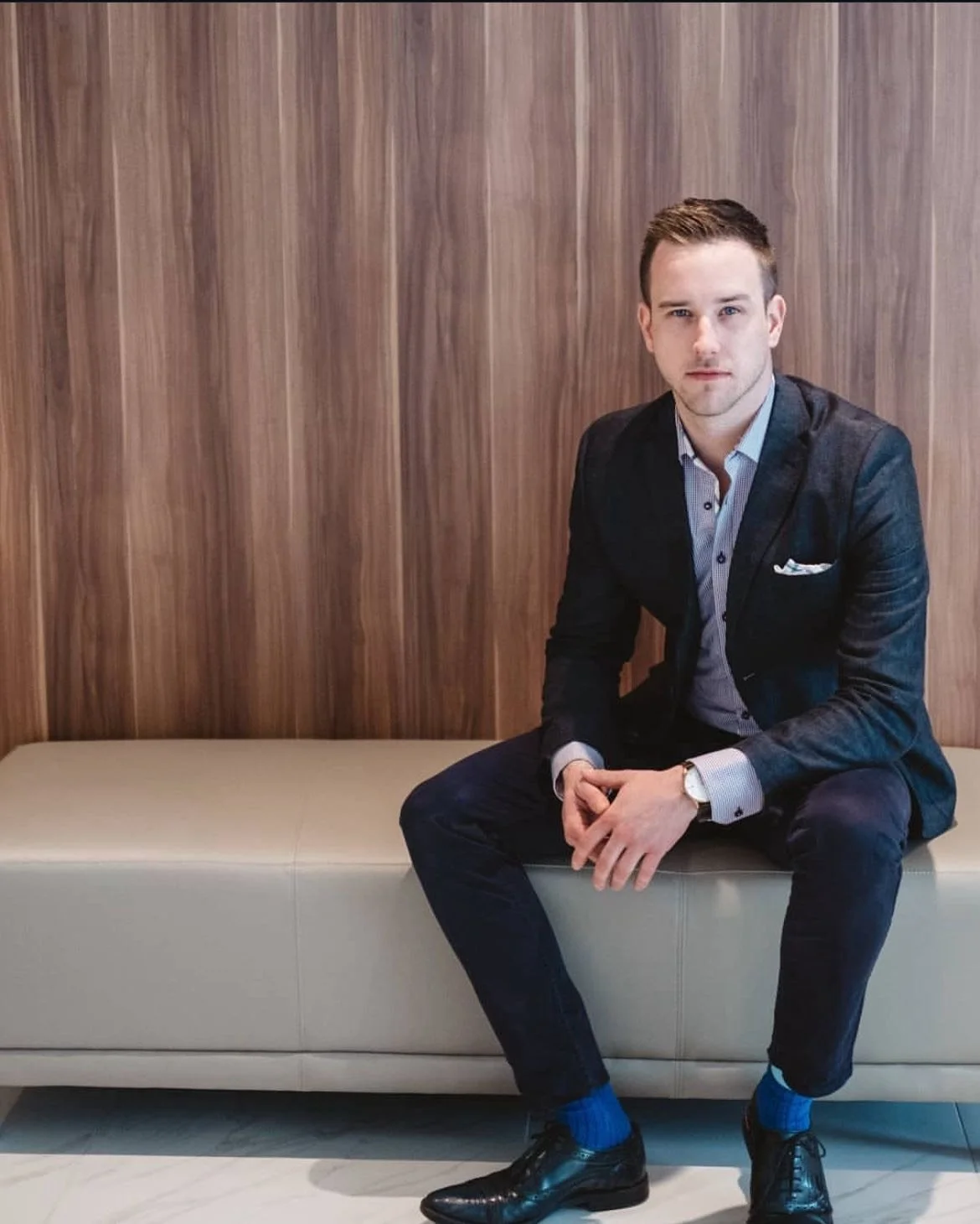 A young man in a business suit sitting on a beige bench with a wooden wall background, wearing a watch, dress shirt, dark pants, and black shoes with blue socks.