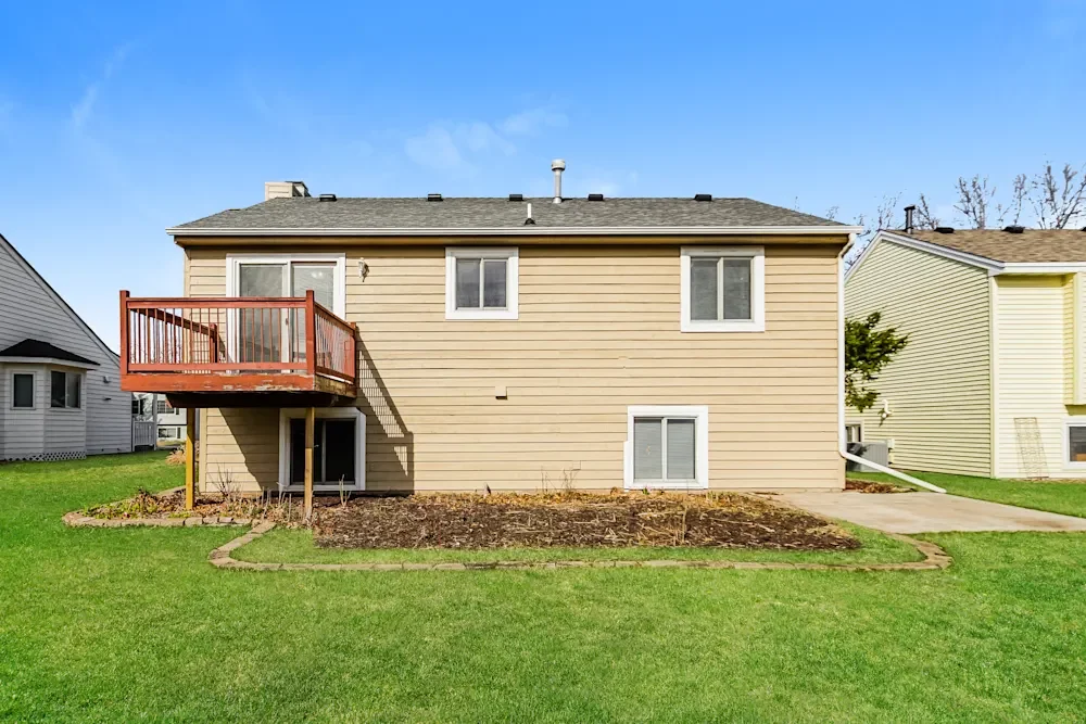 Back view of a two-story beige house with a small wooden balcony on the left side, a sliding door leading to the balcony, four windows, a green lawn, a garden bed, and a concrete pathway.