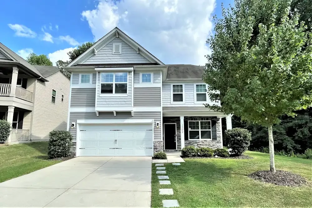 Front view of a two-story house with a white garage door, a small front porch, a tree on the right, and landscaped yard with shrubs and stepping stones.