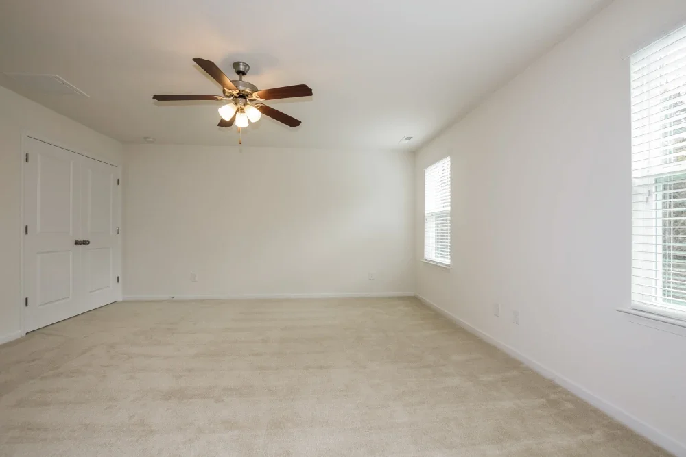 Empty room with white walls, beige carpet, two windows with blinds, a ceiling fan with lights, and closed closet doors.