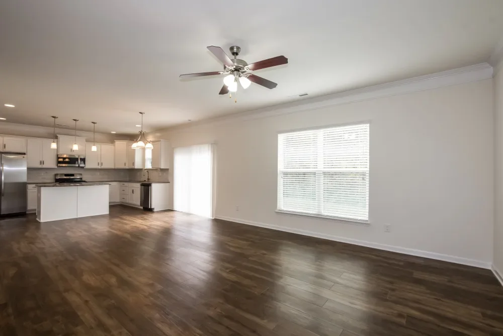 Empty living room with wooden floors, white walls, a ceiling fan, large window with blinds, and open kitchen with white cabinets and stainless steel appliances.