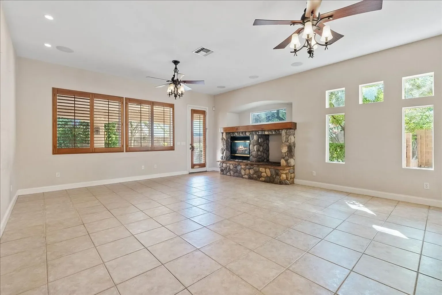 Empty living room with tiled floor, stone fireplace, large window with wooden blinds, ceiling fans, and a glass door with bamboo blinds.