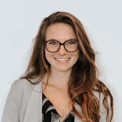 A woman with long brown hair and glasses smiling at the camera against a plain white background.