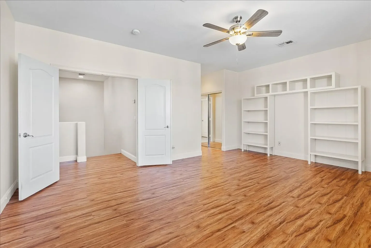 Empty room with white walls, hardwood flooring, built-in bookshelves, ceiling fan, and an open closet door.