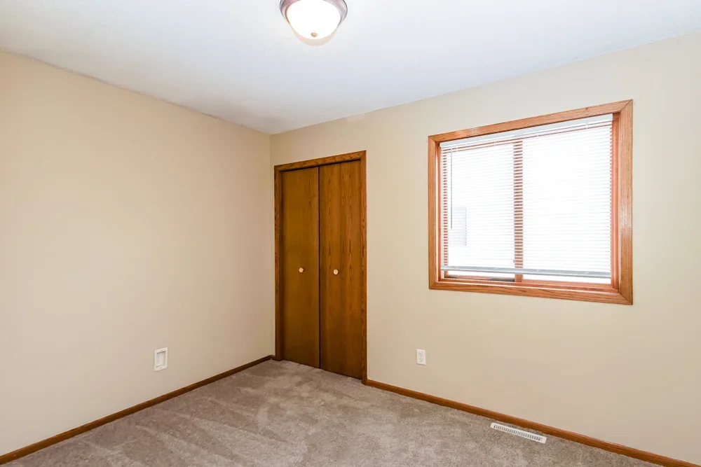 Empty room with beige walls, a window with blinds, wooden trim, a closet with wooden doors, and a ceiling light fixture.