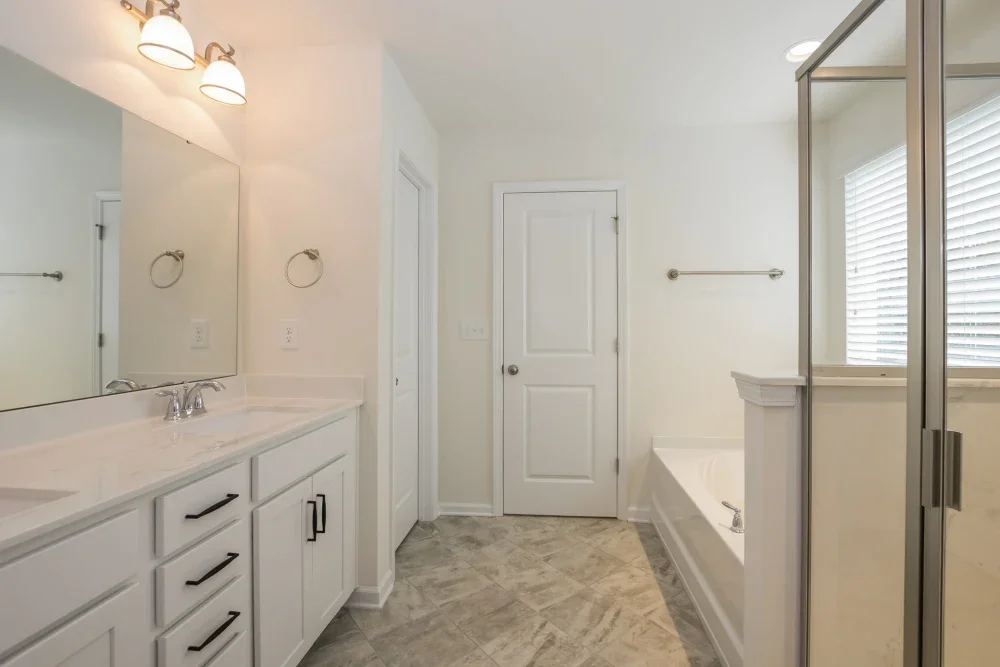 Bathroom with white cabinets, a large mirror, two light fixtures, a bathtub, a glass shower enclosure, and a window with blinds.
