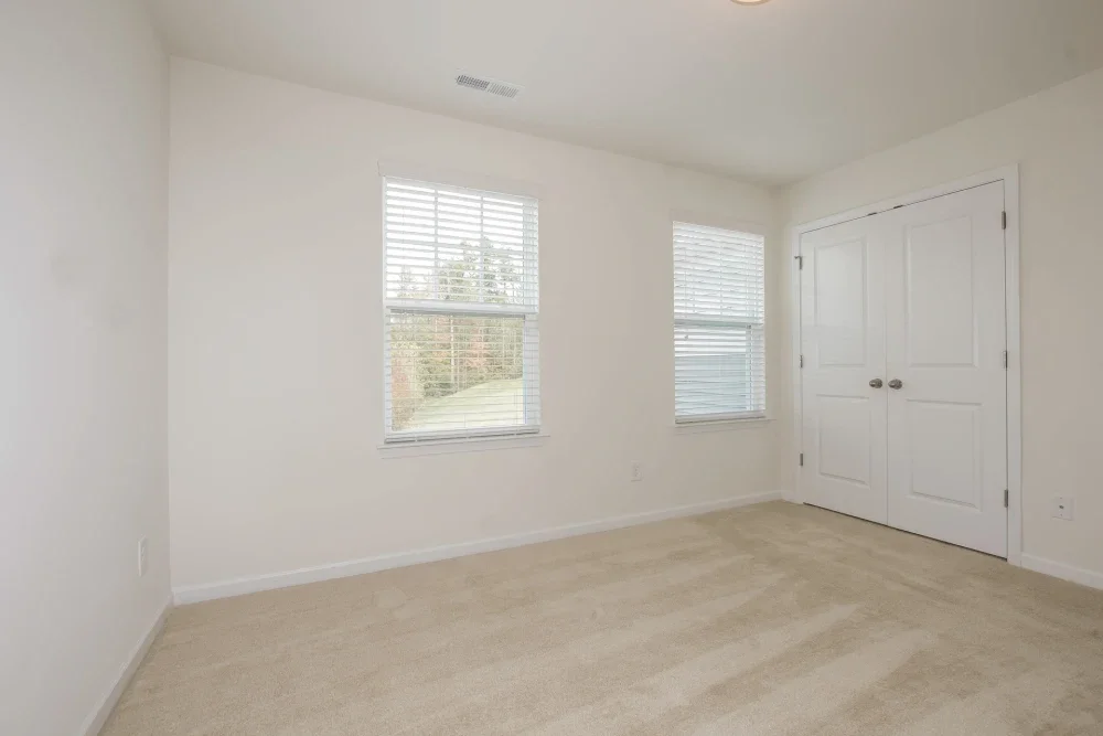 Empty bedroom with white walls, beige carpet, two windows with blinds, and a closed double door closet.