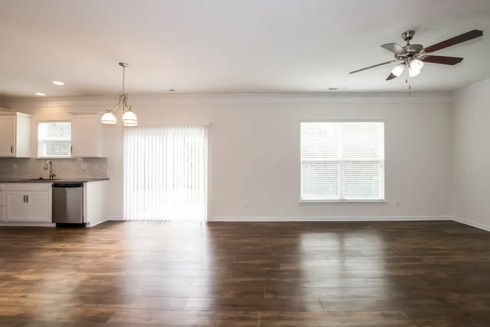 Empty living room with wooden floors, white walls, a ceiling fan, a window, and a sliding glass door leading to a patio.