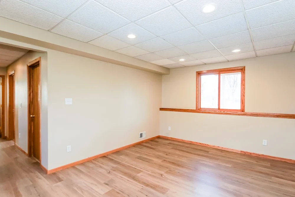 Empty room with beige walls, wooden flooring, and a single window with wooden trim, ceiling lights, and doorways in the background.