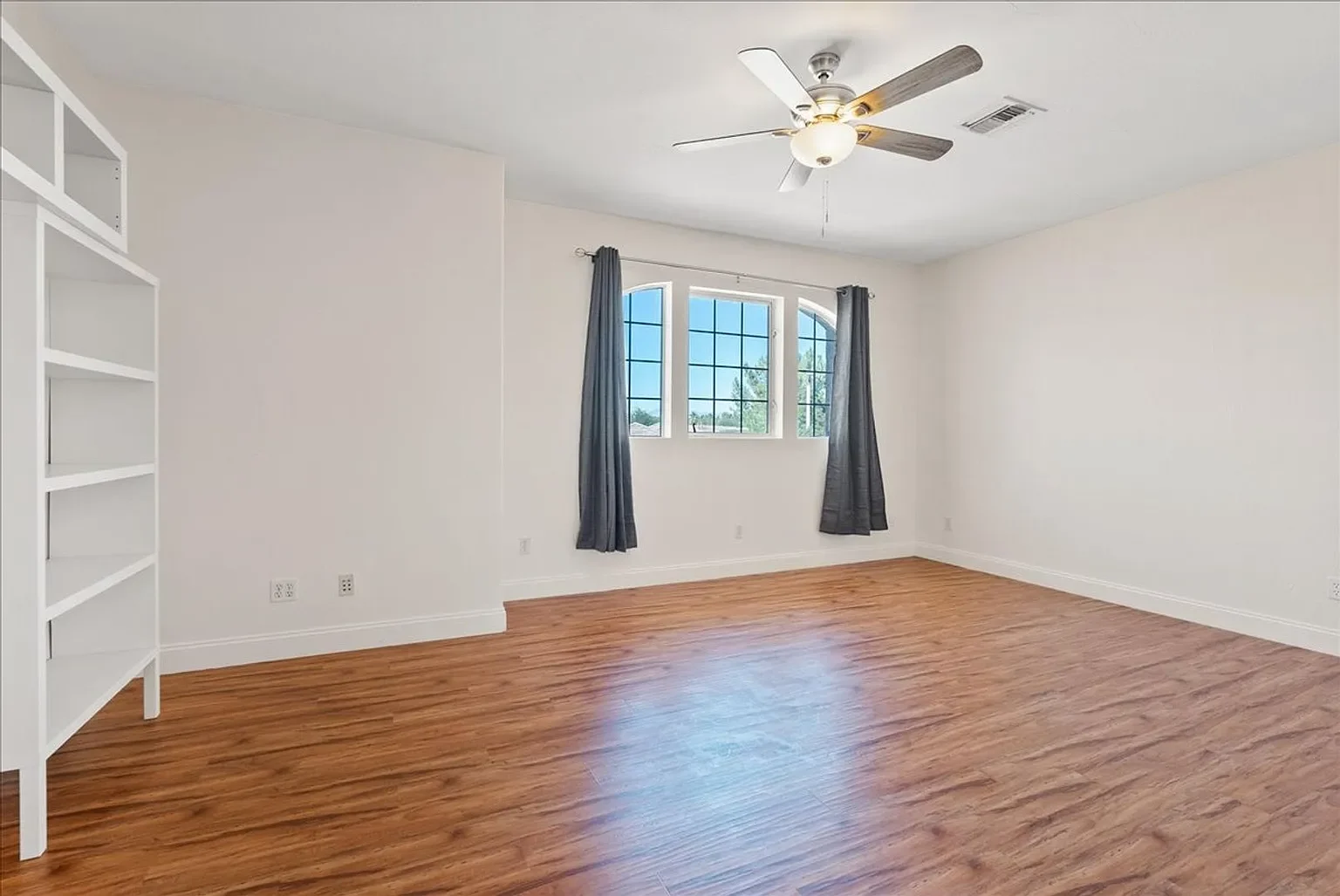 Empty room with white walls, wooden flooring, a ceiling fan, and a window with dark curtains.
