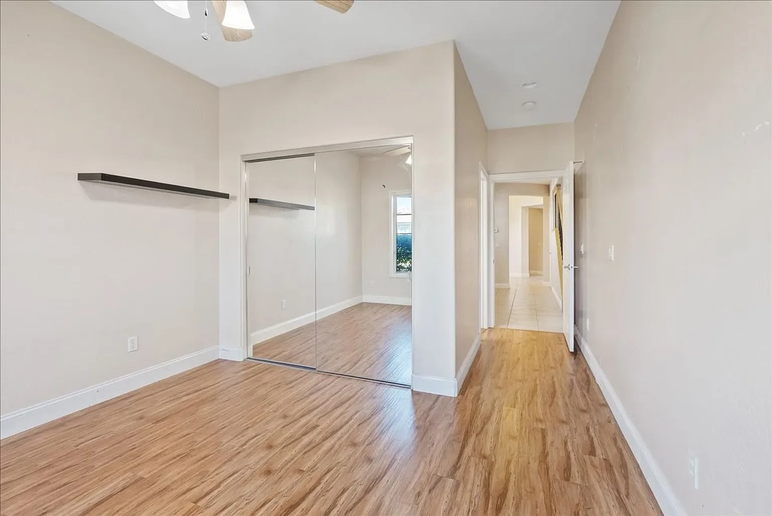 Empty room with hardwood flooring, white walls, a mirrored closet door, and a window with a view outside.
