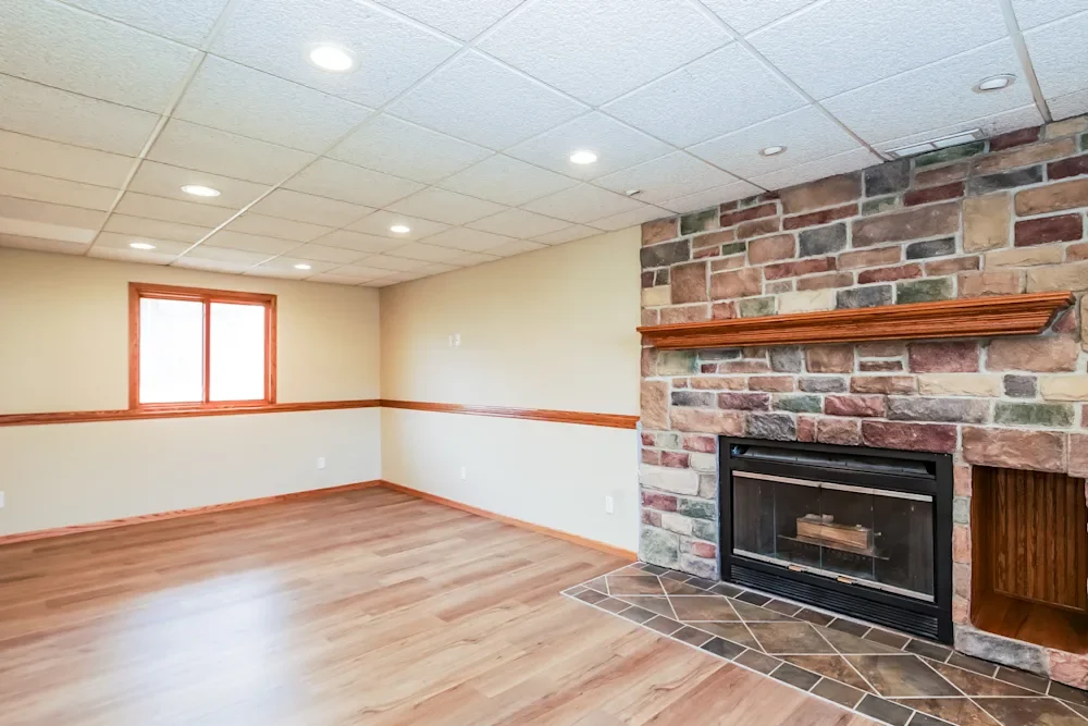 Empty living room with hardwood floors, beige walls with wood trim, a brick fireplace with wooden mantle, and a window on the wall.
