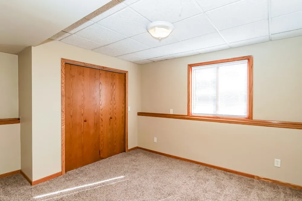 Empty room with beige walls, wooden trim, a window with blinds, a closet with wooden doors, and a ceiling light.