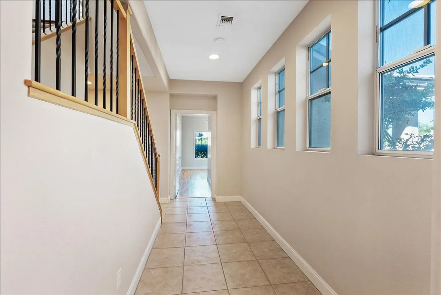 Bright hallway with tiled floor, multiple windows on right side, white walls, and a staircase with black railings on the left, leading upstairs.