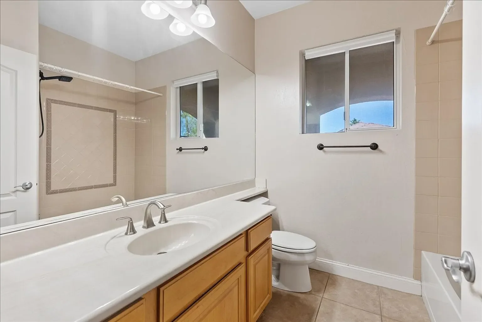Bathroom with a large mirror above a countertop with a sink, a toilet, two small windows, a shower with tiled walls, and a beige tiled floor.