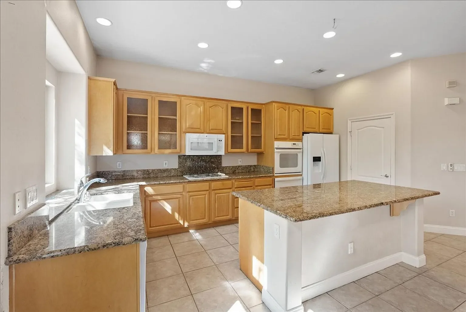 Empty kitchen with wooden cabinets, granite countertops, white appliances, and a kitchen island.