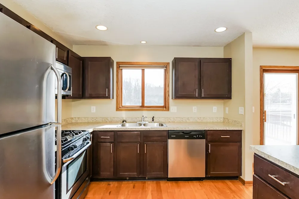 Kitchen with stainless steel refrigerator, microwave, oven, and dishwasher, dark wood cabinets, beige countertops, and a window above the sink, with a sliding door on the right.