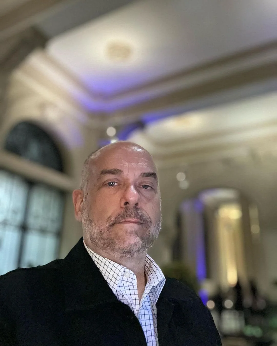 A man with a beard and light-colored hair takes a selfie in an elegant, high-ceilinged room with architectural details and soft lighting.