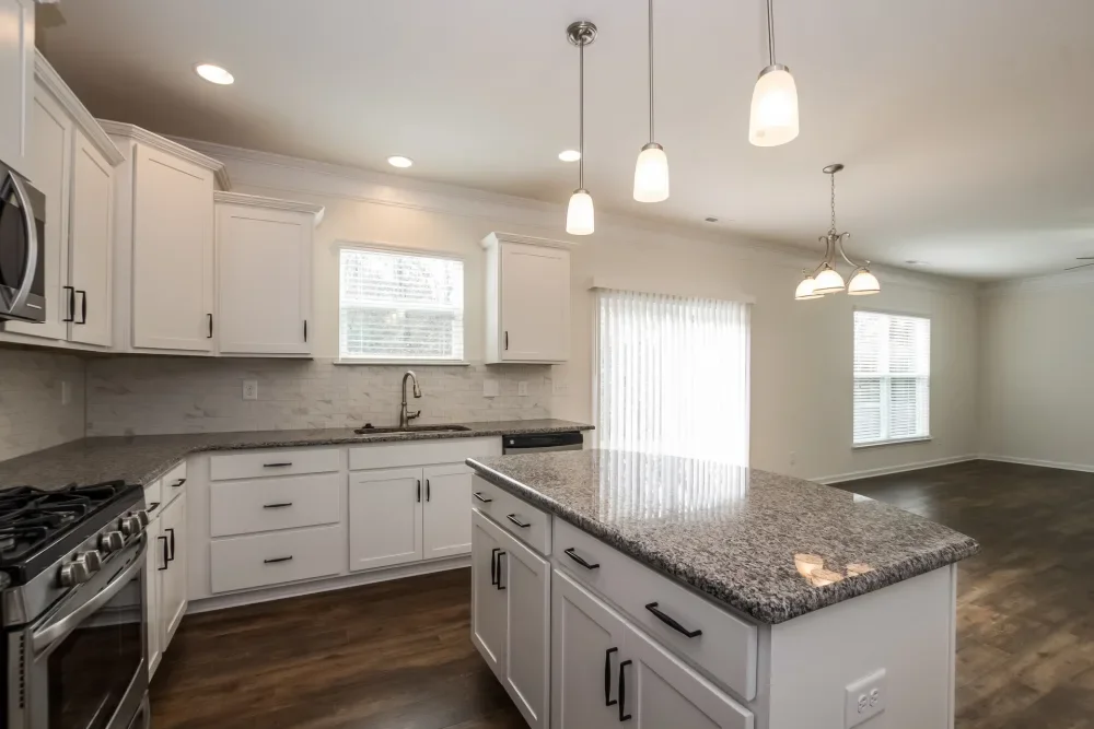 Empty kitchen with white cabinets, granite countertops, stainless steel appliances, wood flooring, and multiple windows, with a view of an open living space.