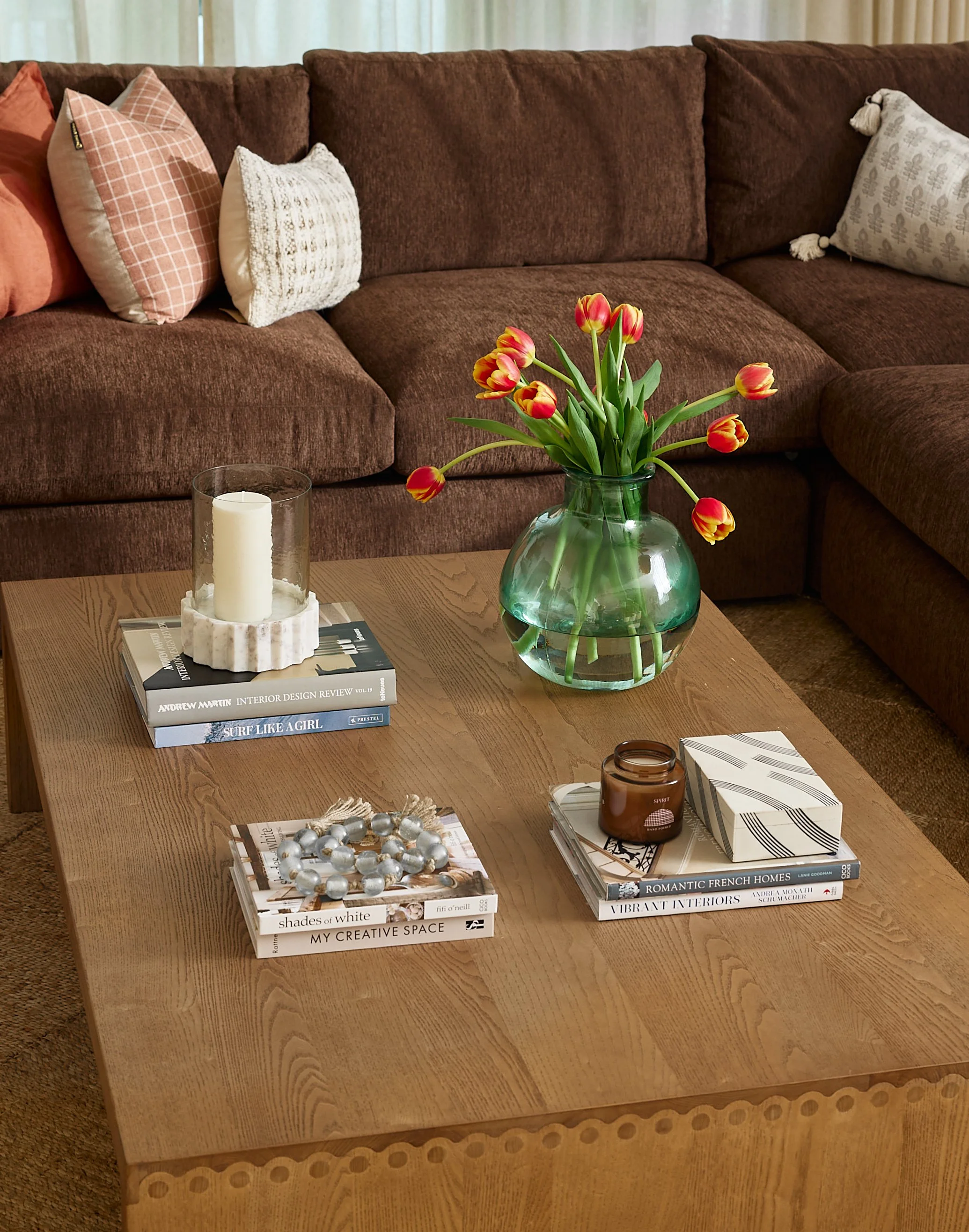 Living room with a brown sectional sofa, colorful throw pillows, a wooden coffee table with a glass vase of tulips, books, a candle, and a decorative bead necklace.