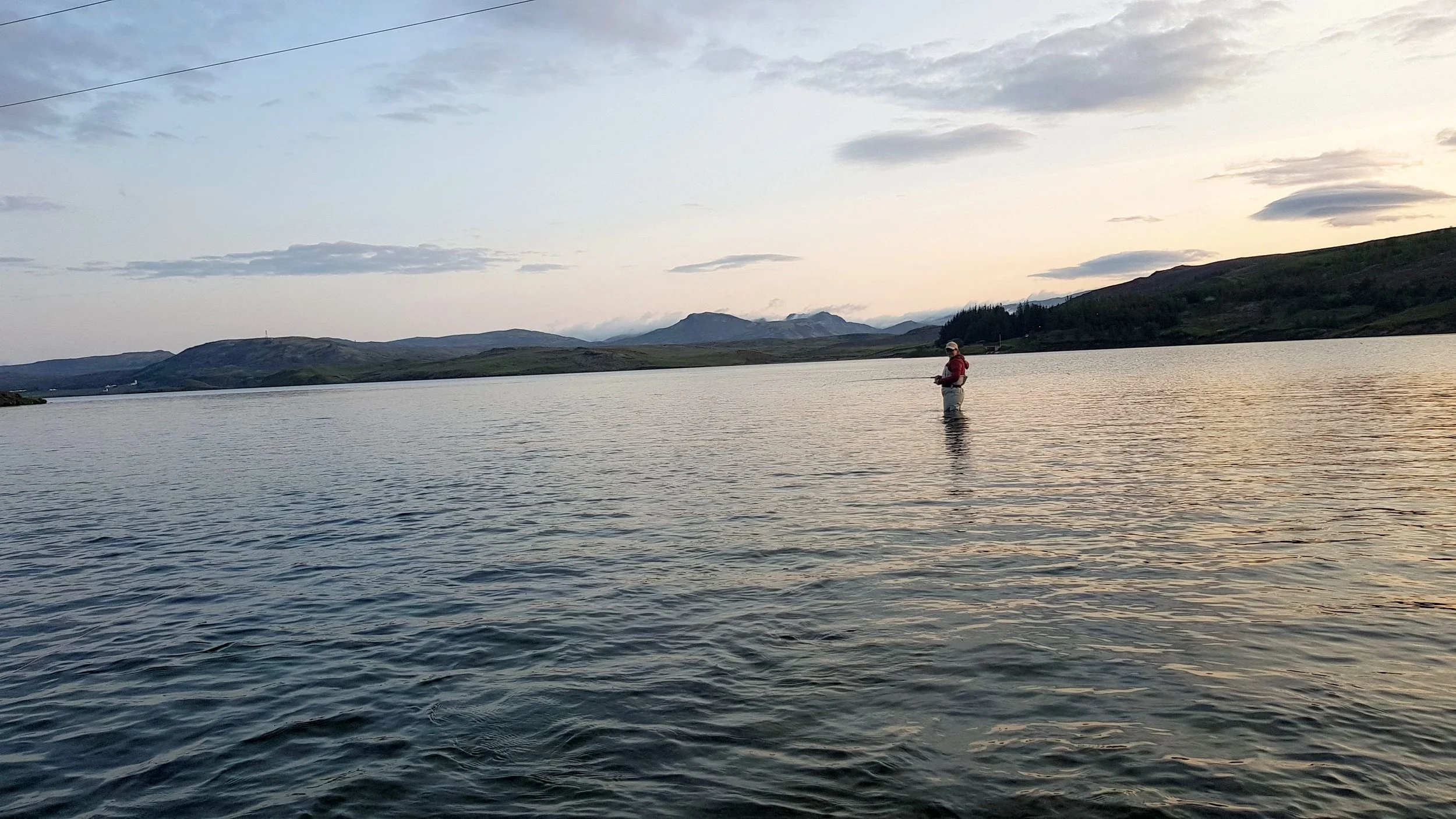 A person stands in a lake or river, fishing, with hills and mountains in the background during a sunset or sunrise.