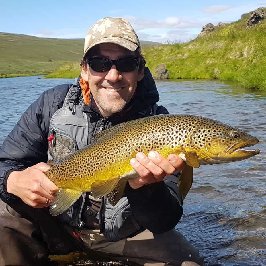 Smiling man in outdoor gear holding a large brown trout near a river with green hills in the background.