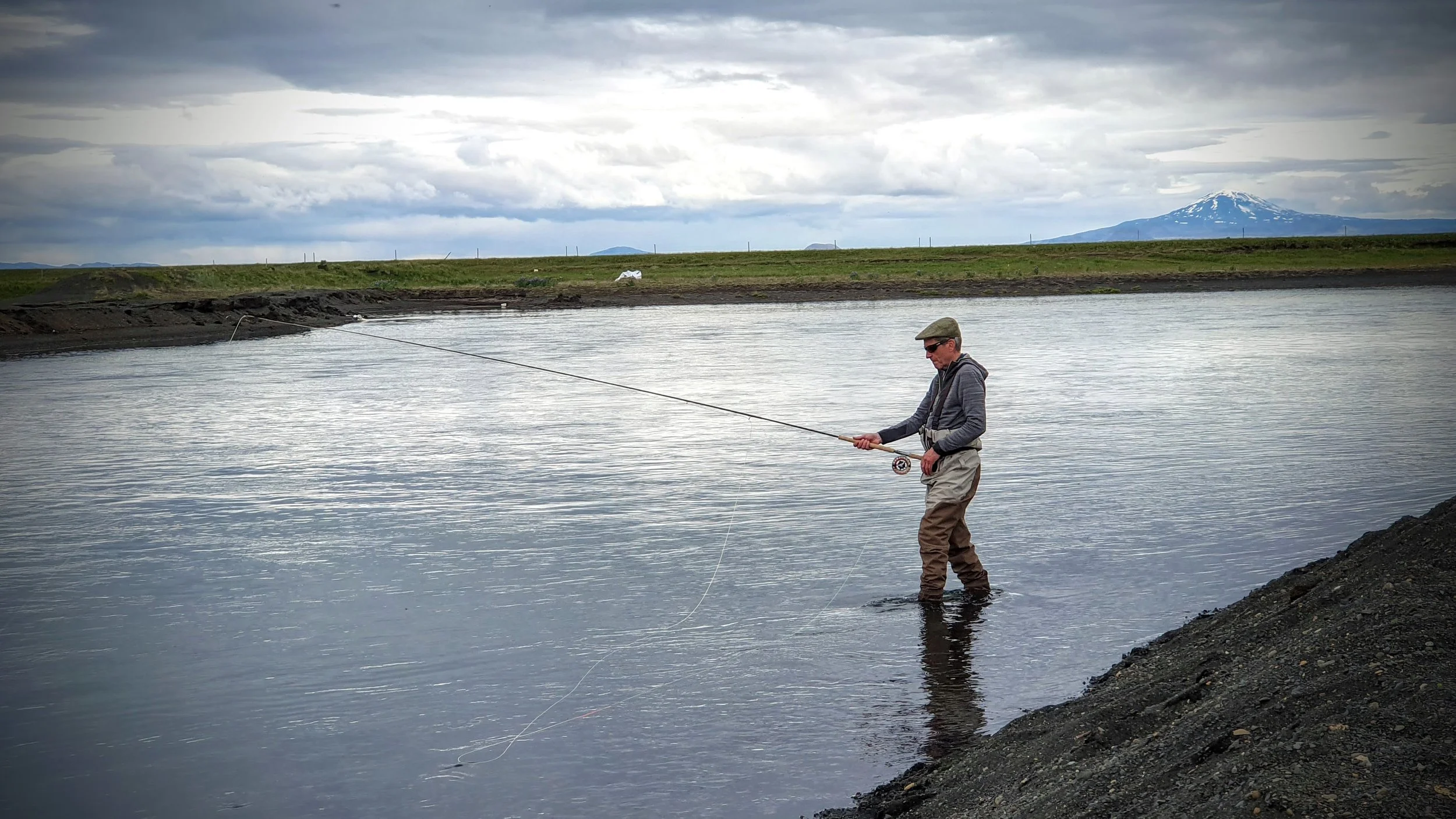 A man in waders standing in a body of water, fishing with a rod in a landscape with green grass, cloudy skies, and a mountain with snow in the background.