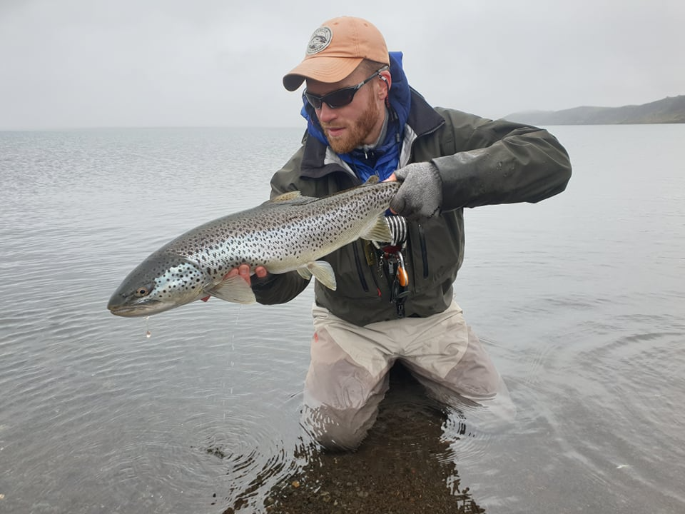 Man in waders holding a large fish in shallow water with a distant shoreline in the background.
