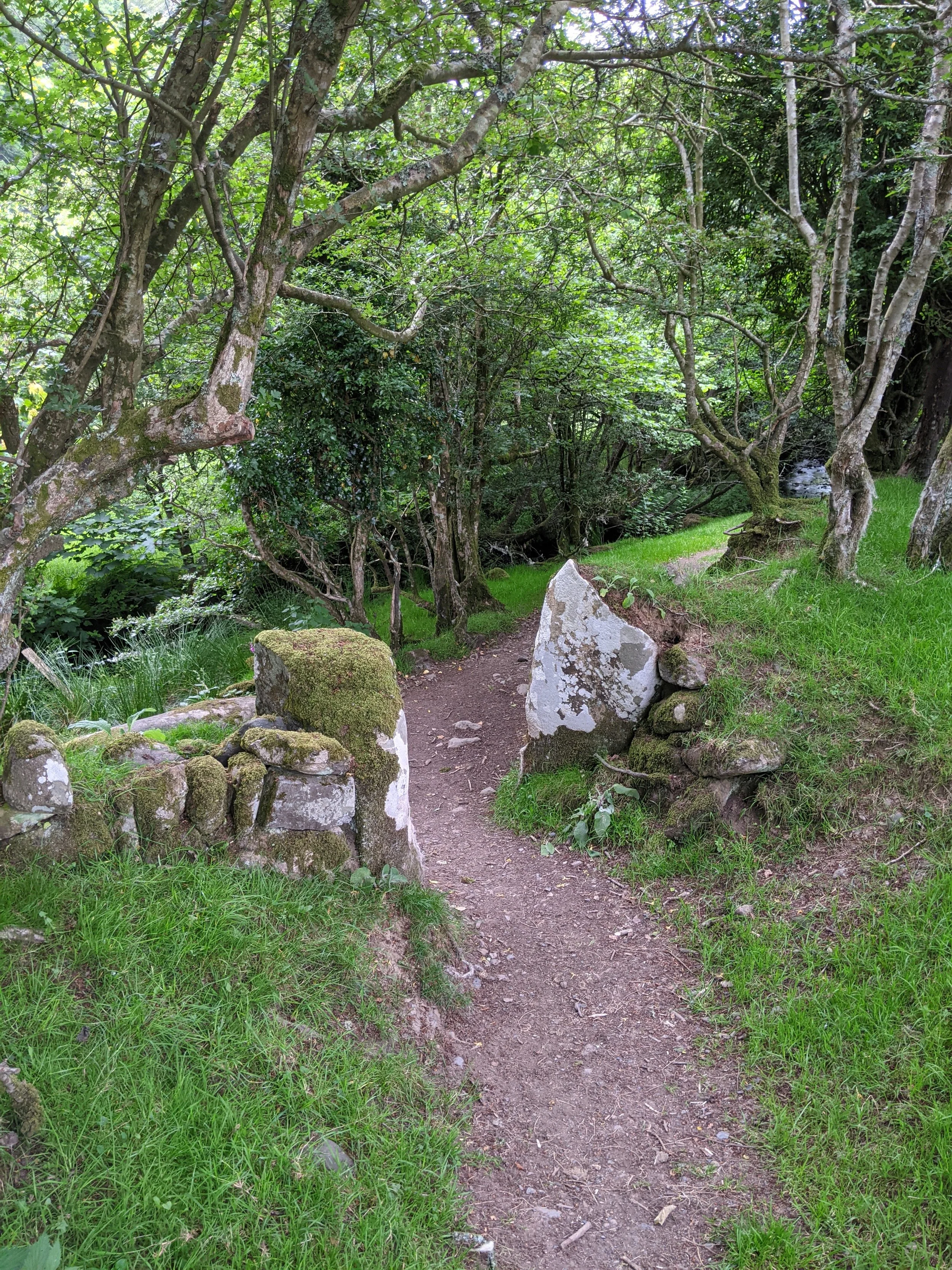 A narrow dirt trail through a lush green forest with moss-covered Stonehenge-style rocks arranged as a gate at the entrance.
