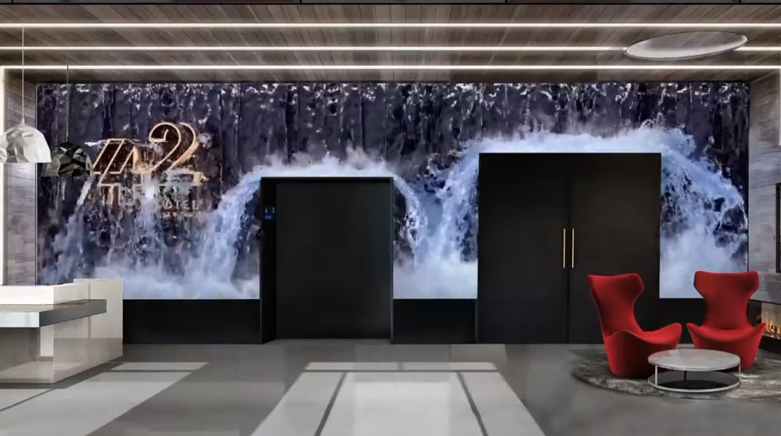 Modern hotel lobby with a water-themed mural on the back wall, featuring a waterfall, a black reception desk, and two red lounge chairs with a round coffee table.